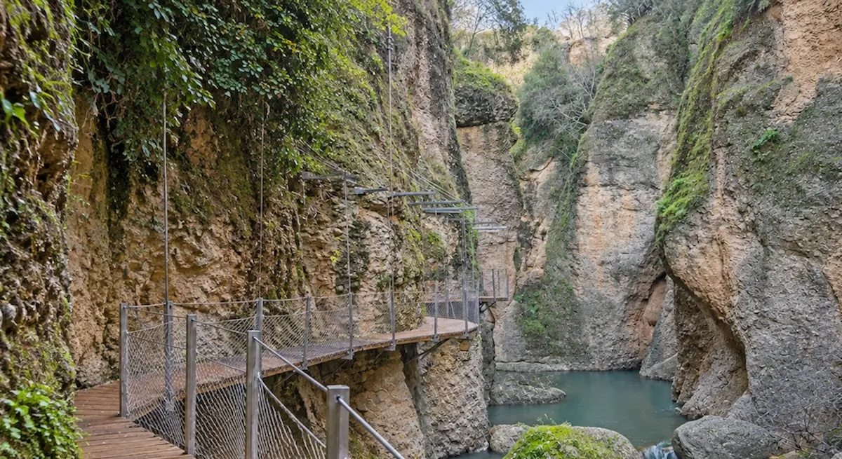 Wooden walkway with a metal railing fixed to the rock wall of the Tajo gorge, above the green water of the río Guadalevín